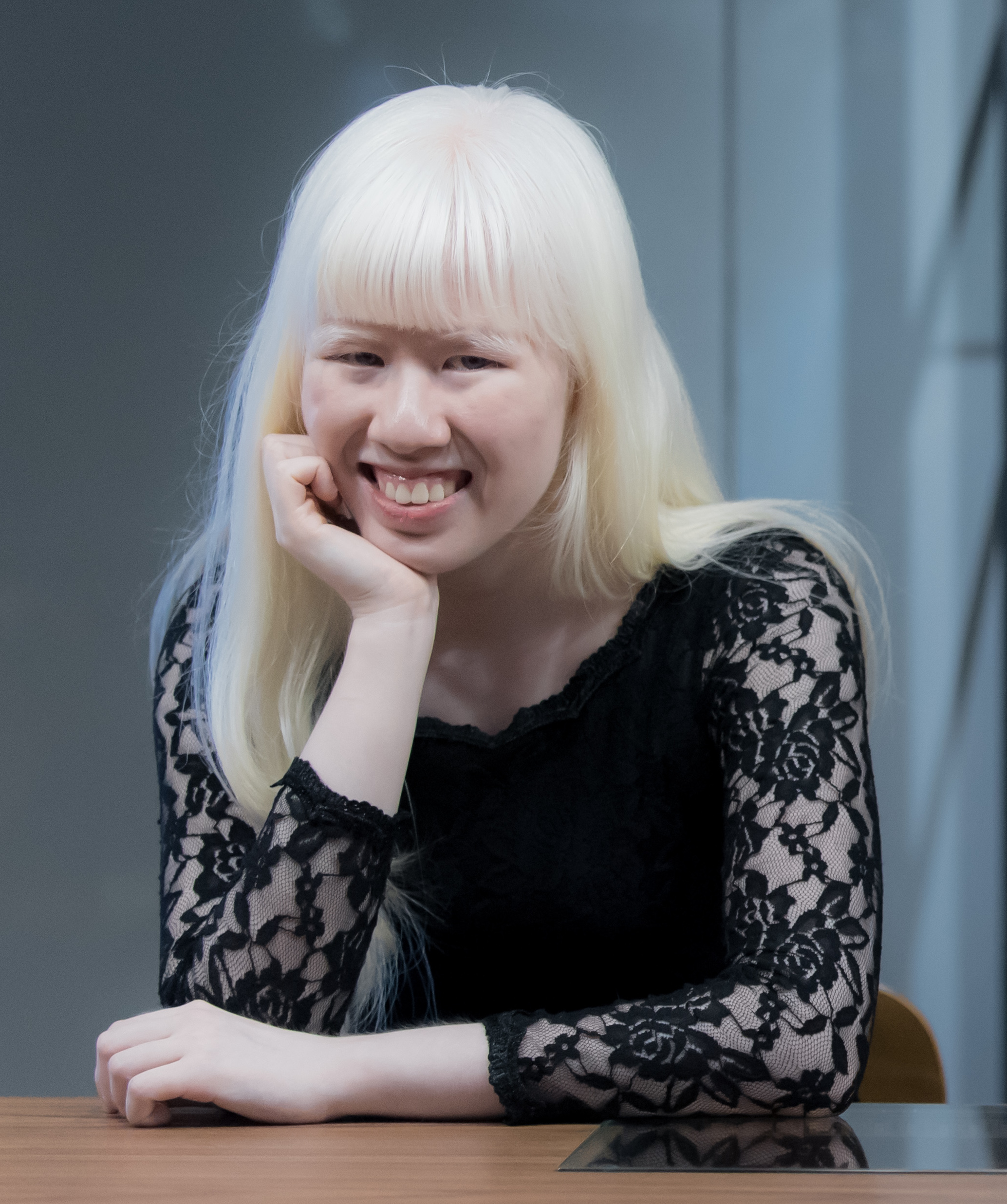 A young person with long white hair smiles warmly, resting their chin on their hand. They wear a black lace top, seated at a wooden table indoors.