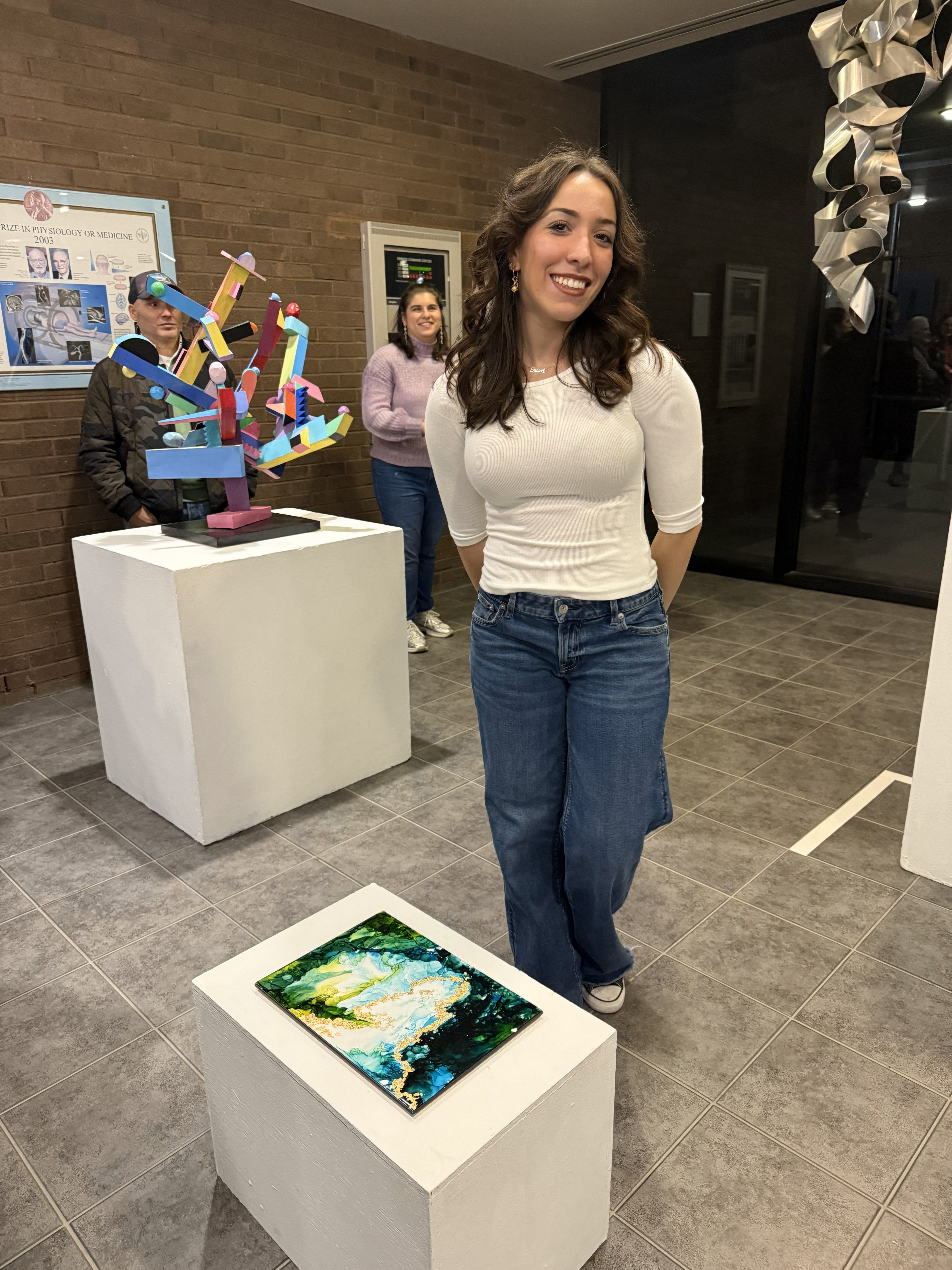 Art student, Eleni Tsevis wearing a white shirt and jeans stands next to her green and gold acryclic art sculpture