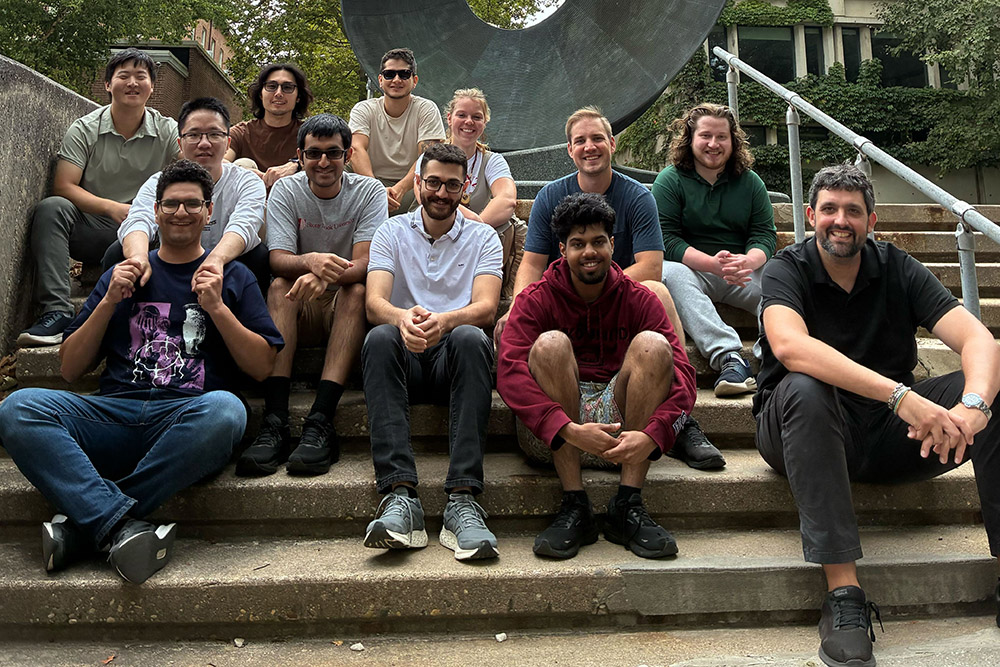 Group of eleven individuals From The Stony Brook University Department of Physics and Astronomy posing on steps outdoors, smiling, with a large sculpture in the background.