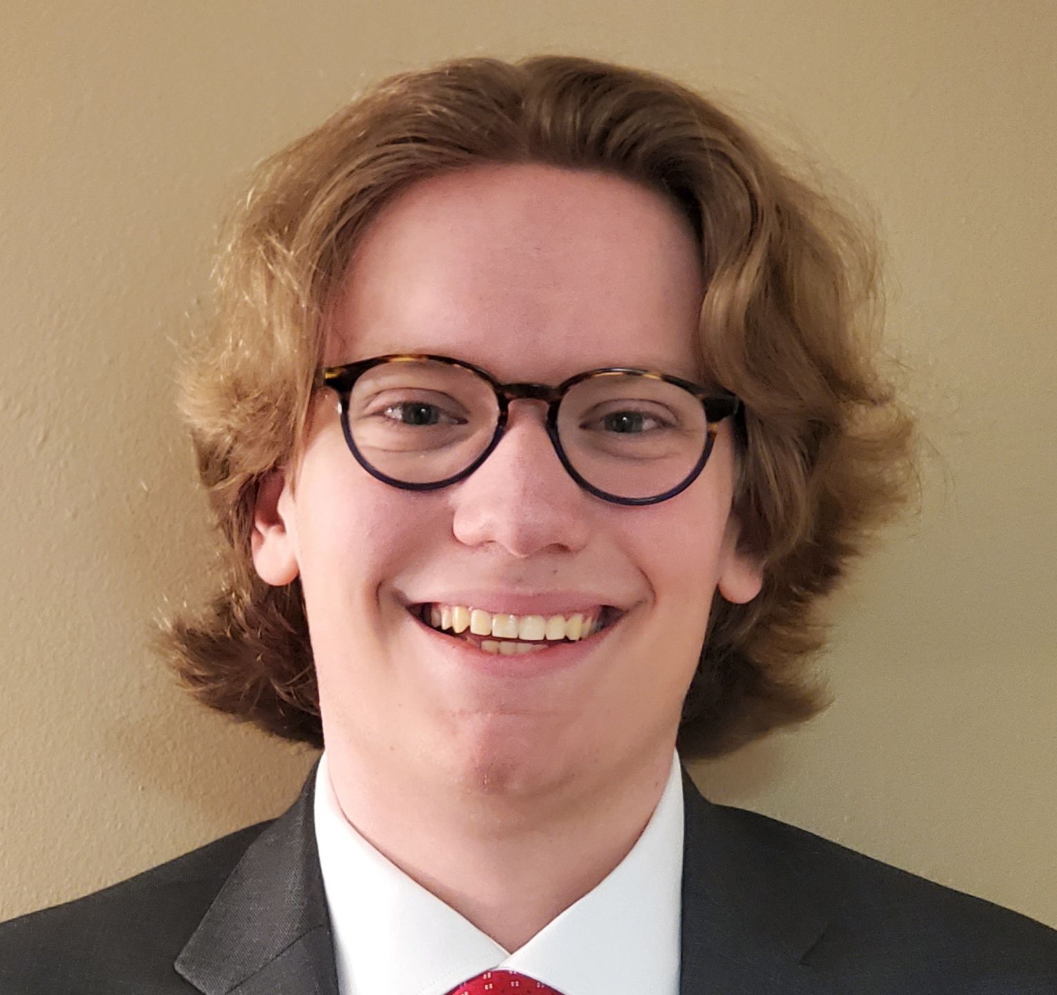 Portrait of a smiling person with shoulder-length curly hair, wearing glasses, a red tie, and a dark jacket.