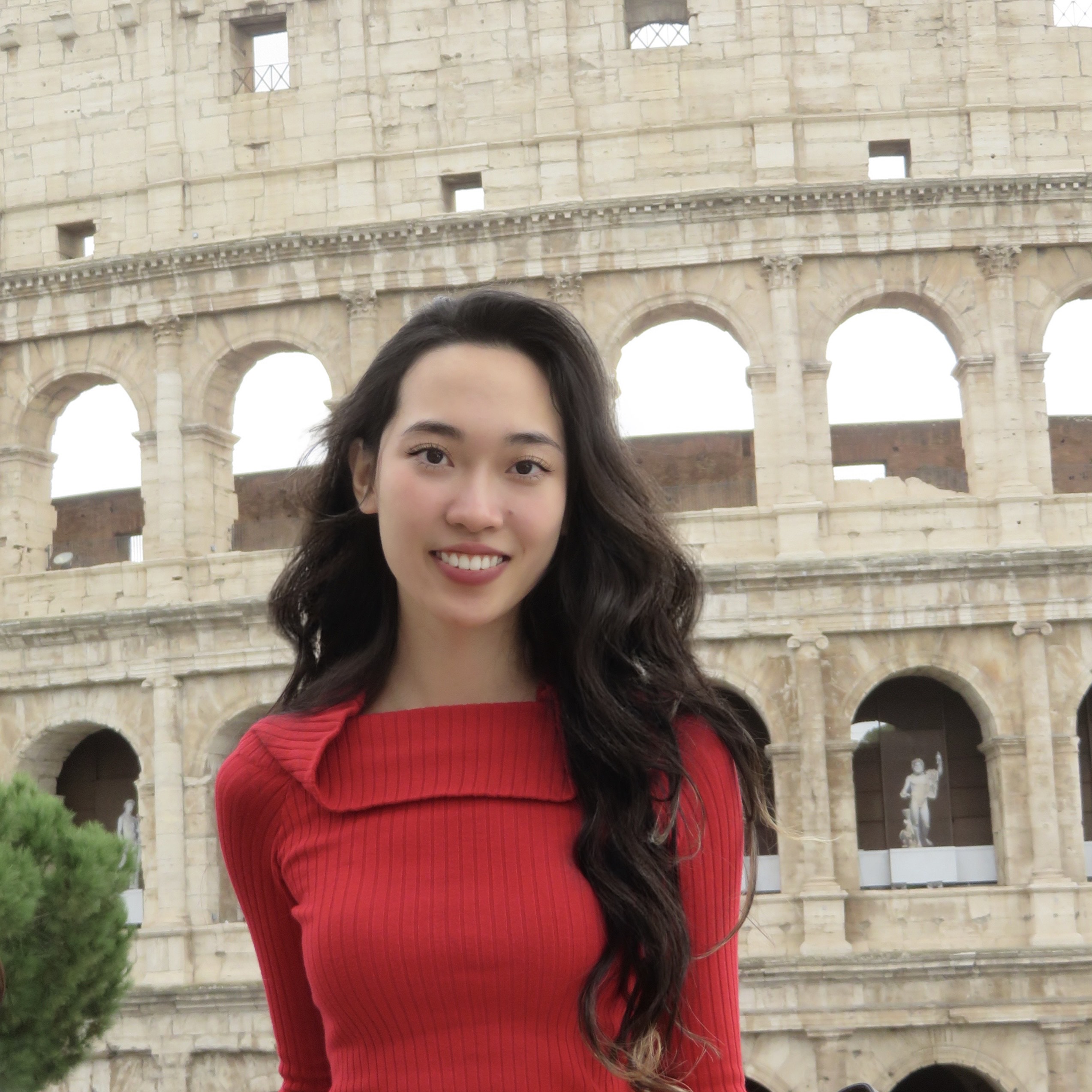 A smiling woman in a red sweater stands in front of the Colosseum in Rome. The ancient structure's arches create a historic backdrop, conveying travel joy.