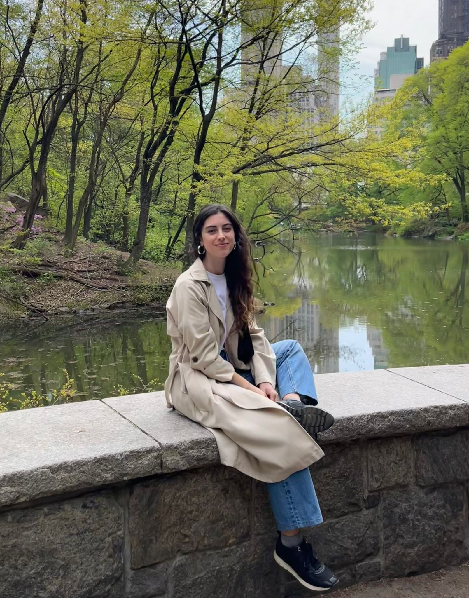 Person sitting on a stone wall in Central Park with a pond and lush greenery in the background.