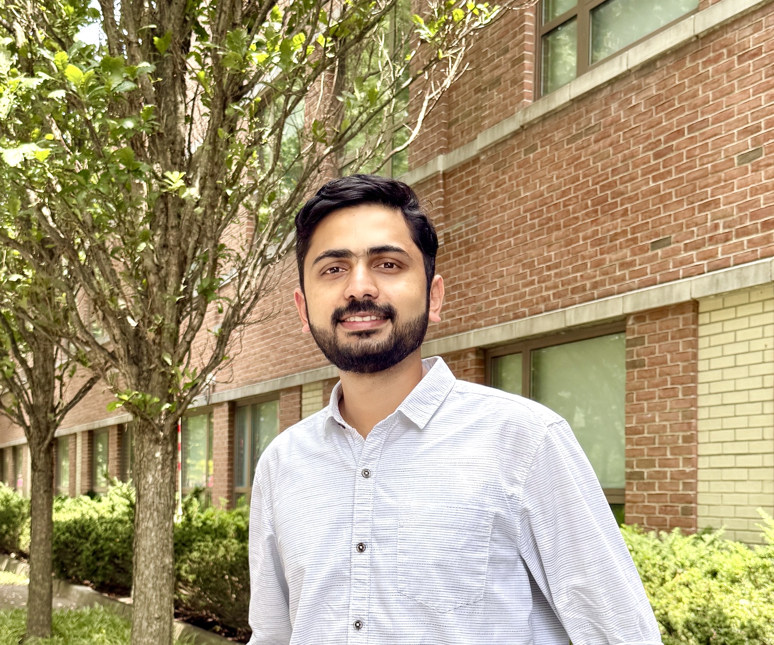 Person in a light blue shirt smiling and standing outdoors in front of brick buildings and green trees.