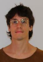 Portrait of an individual with shoulder-length brown hair and round glasses, wearing a brown shirt, against a light background.