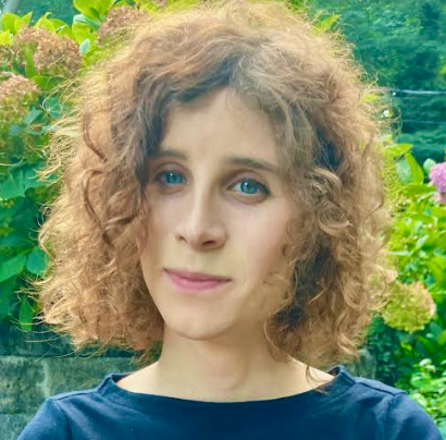 A person with curly hair standing in front of green foliage.