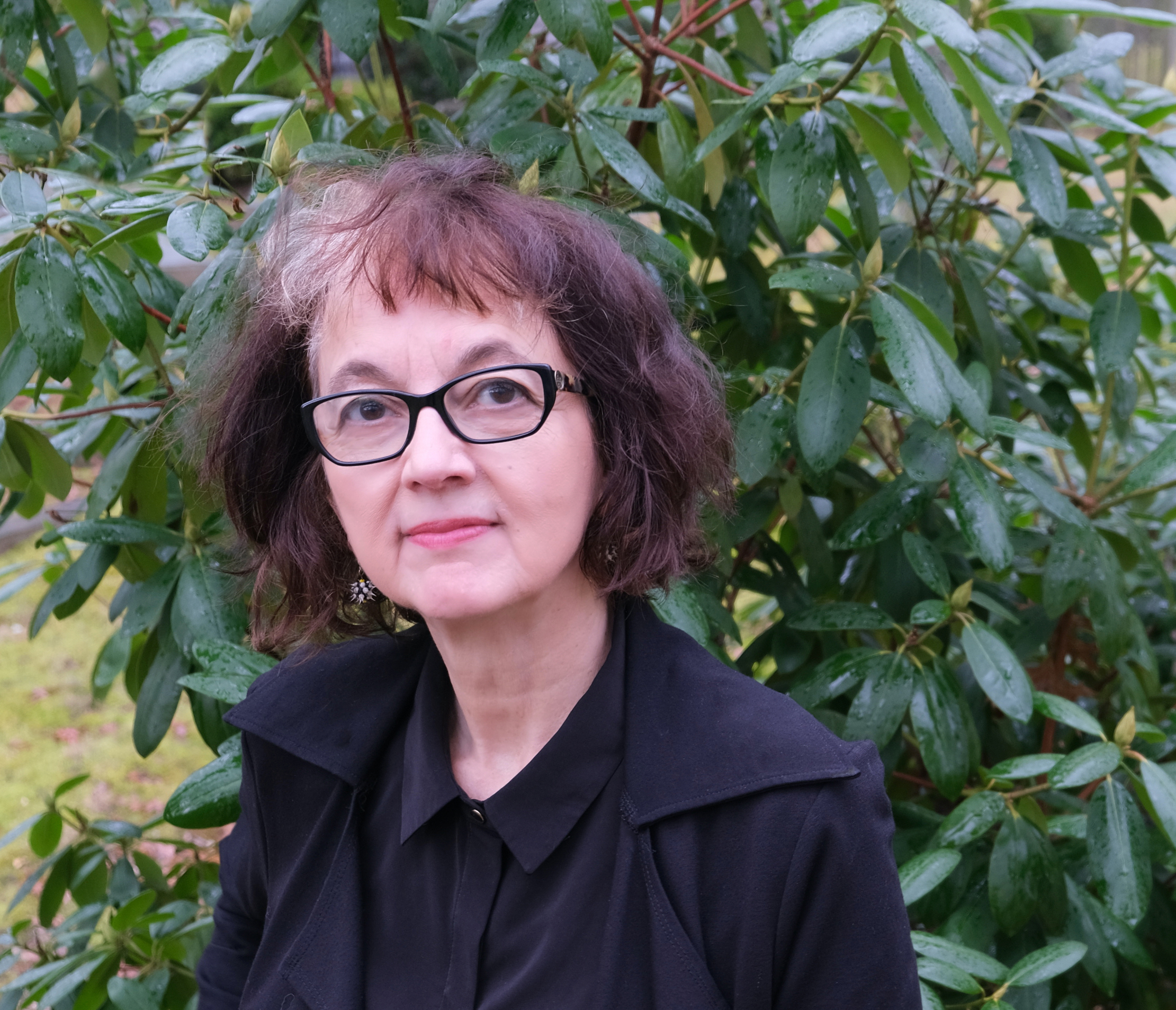 Portrait of an individual with short curly hair and glasses, wearing a black outfit, posing in front of a lush green bush.