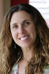 Portrait of a smiling person with long, curly hair wearing earrings and a floral top, standing by a window.