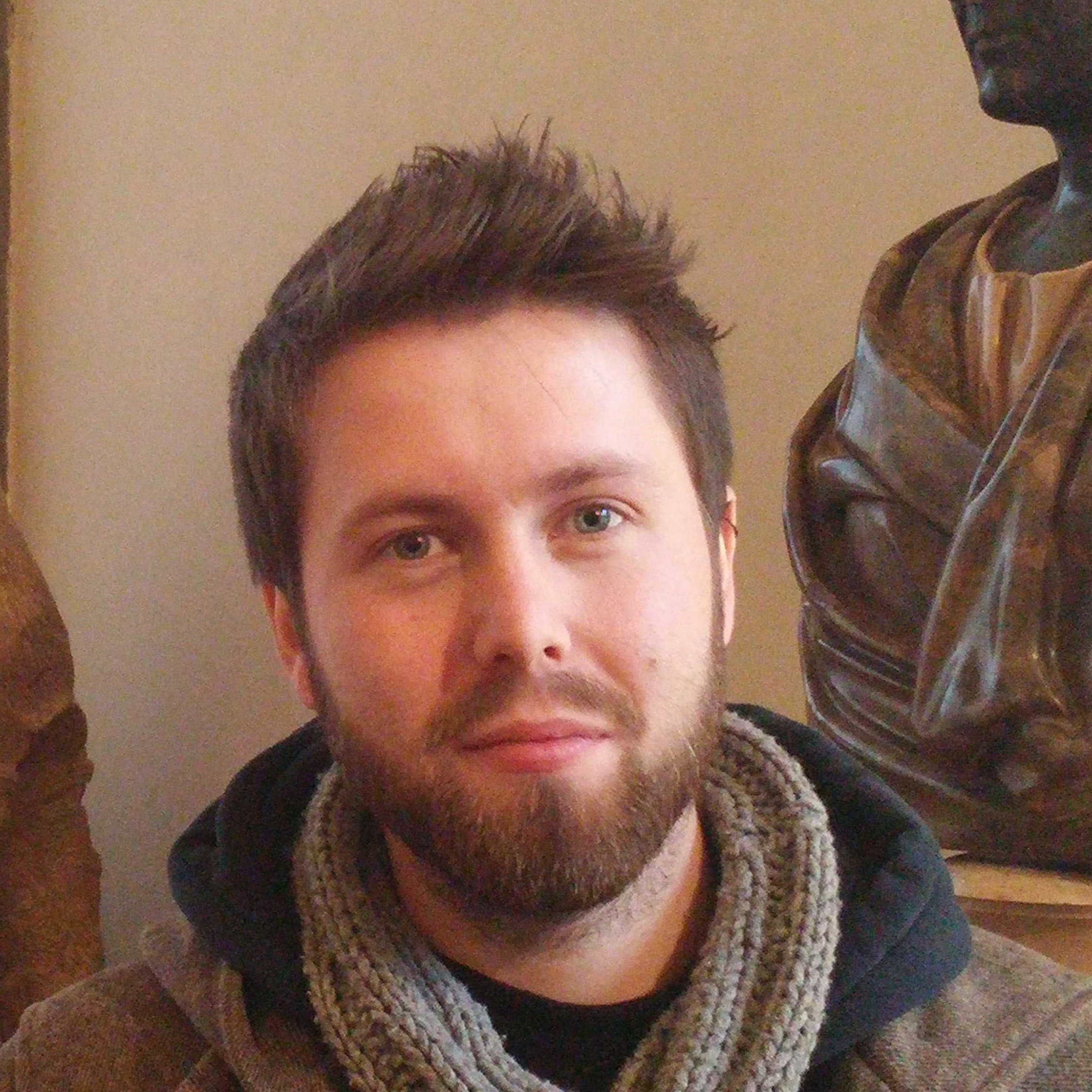 Person with a short beard and tousled hair, wearing a gray scarf, sitting in front of a classical sculpture.