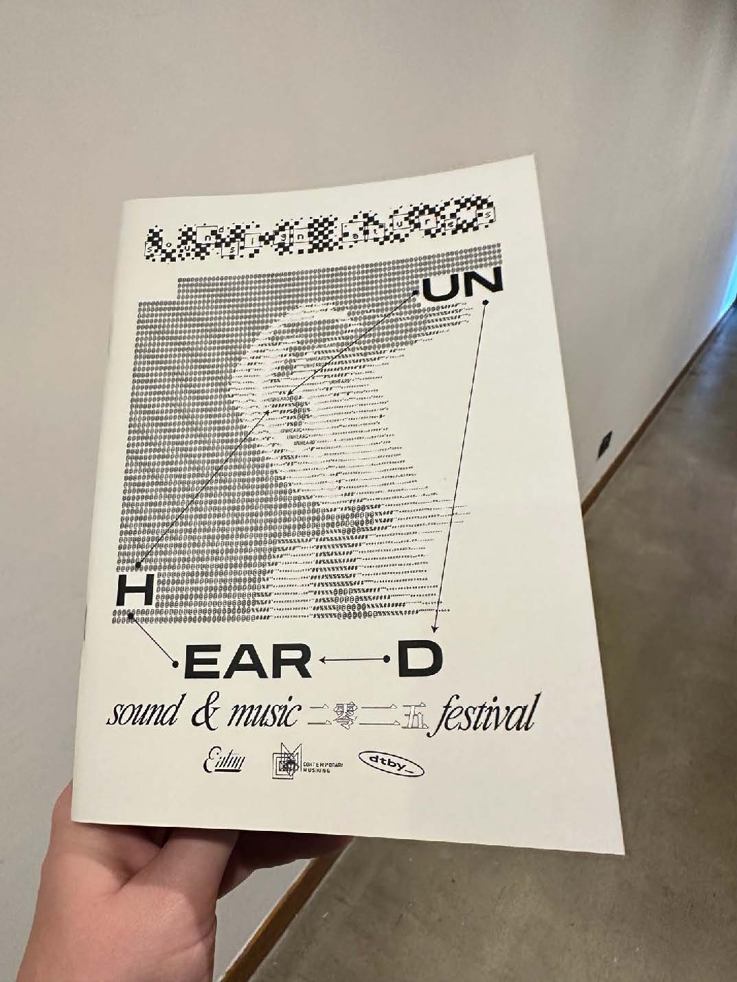 A hand holding a promotional flyer for the "Unheard Sound & Music Festival," featuring a stylized pixelated graphic of an ear and the UN logo. The flyer is predominantly black and white with text and various small symbols related to sound and music.