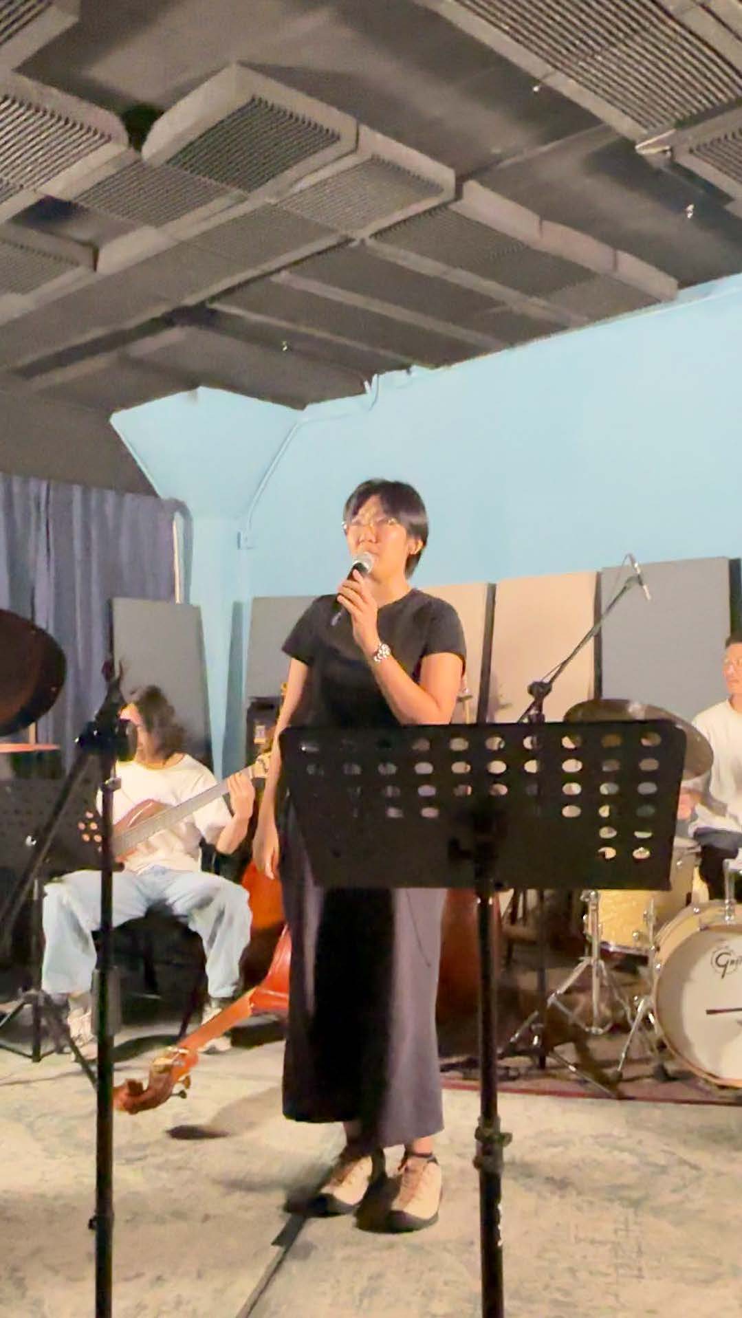 Music performance Person singing into a microphone with a band performing in the background in a room with overhead ductwork.