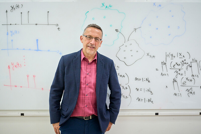 Electrical and Computer Engineering Faculty Man in glasses and a blazer stands confidently in front of a whiteboard filled with mathematical equations and graphs, conveying a scholarly atmosphere.