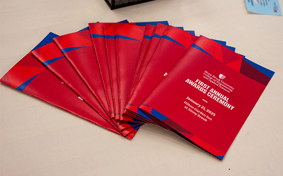 A stack of red and blue event programs for the First Annual Awards Ceremony, held on January 23, 2025, at Stony Brook University, displayed on a table.