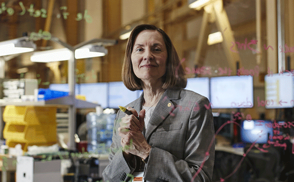 Woman in a gray suit stands confidently in an office filled with computers, writing equations on a transparent board. The atmosphere is focused and analytical.