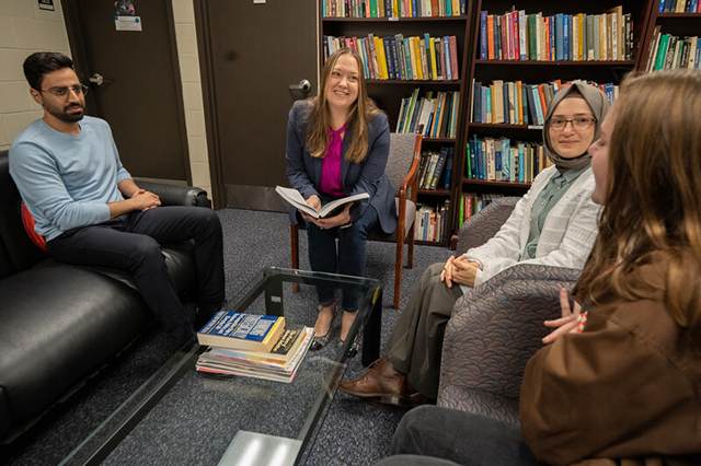 Technology and Society Faculty A group of four people sits in a library on a couch and chairs, engaged in a discussion. Bookshelves are filled in the background, creating a scholarly atmosphere.