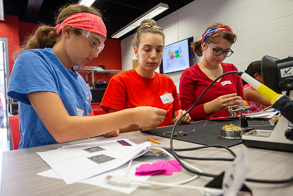 Three young people in safety goggles and casual clothes work intently on electronics at a table in a workshop, reflecting concentration and teamwork.