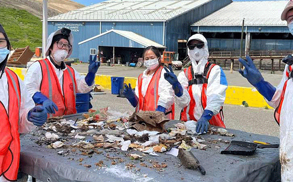 Five people wearing protective gear, vests, gloves, and masks sort trash on a table outdoors. The scene conveys teamwork and environmental cleanup.
