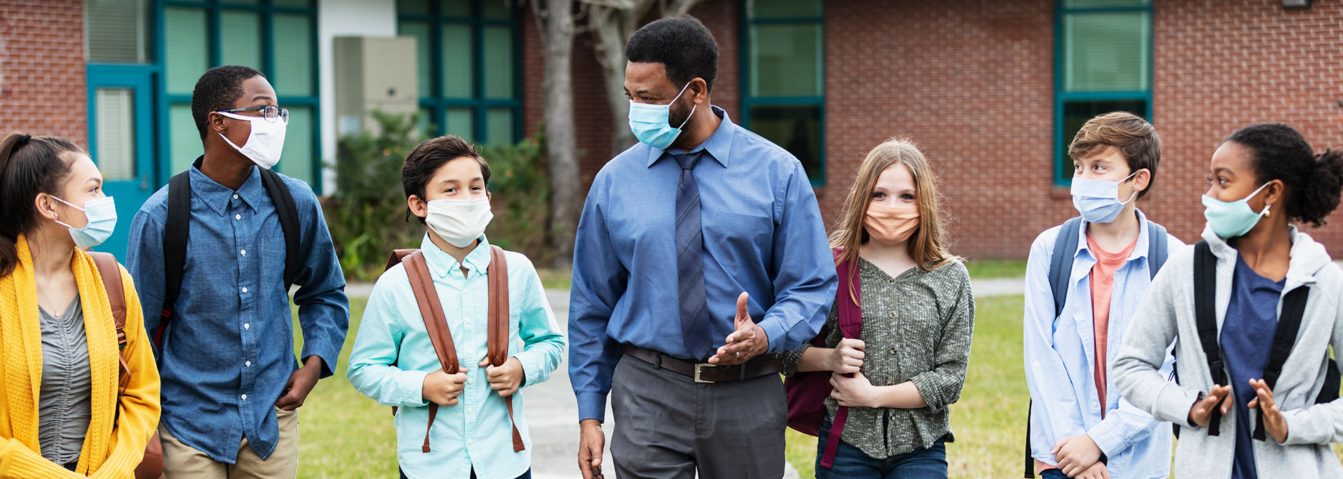 Masked teacher and masked school kids walking in a line in front of a school