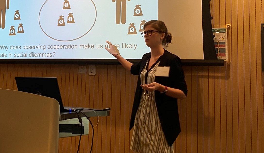 A young woman with glasses and her hair tied back stands in a wood-paneled room, giving a presentation. She is wearing a black blazer over a striped dress and is gesturing toward a projection screen behind her. The screen displays an illustration of people and money bags with the text, "Why does observing cooperation make us more likely to cooperate in social dilemmas?" A laptop is visible on a podium in the foreground.