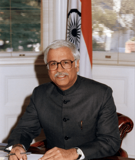 A professional portrait of a man with grey hair, a mustache, and glasses, wearing a grey Nehru-style jacket. He is seated at a wooden desk, holding a pen over a document. In the background, a white wall with wainscoting is visible next to a window with blinds, and the flag of India stands behind him.