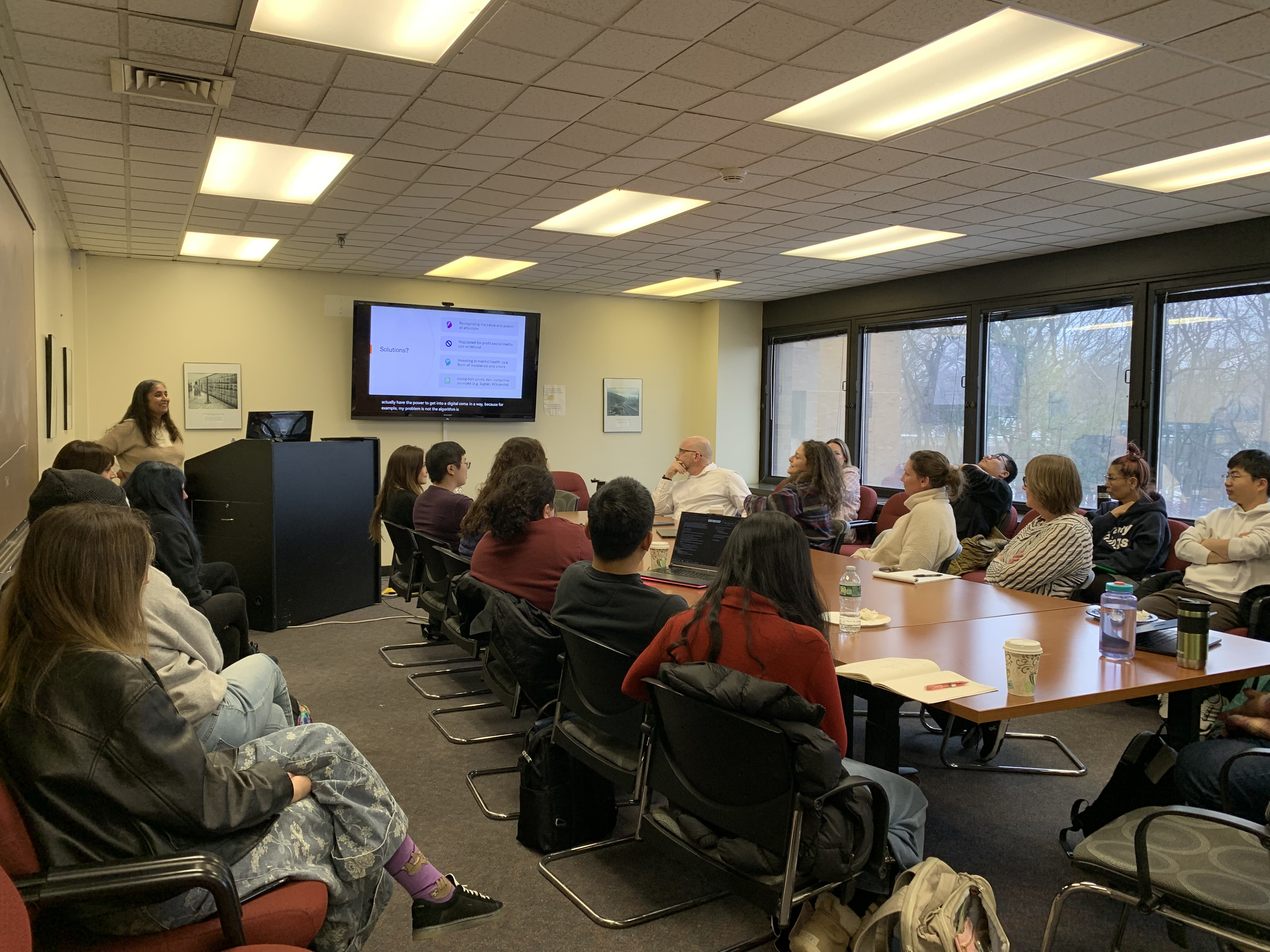 A classroom with a presenter at a podium and an audience seated, viewing a presentation on a large screen.