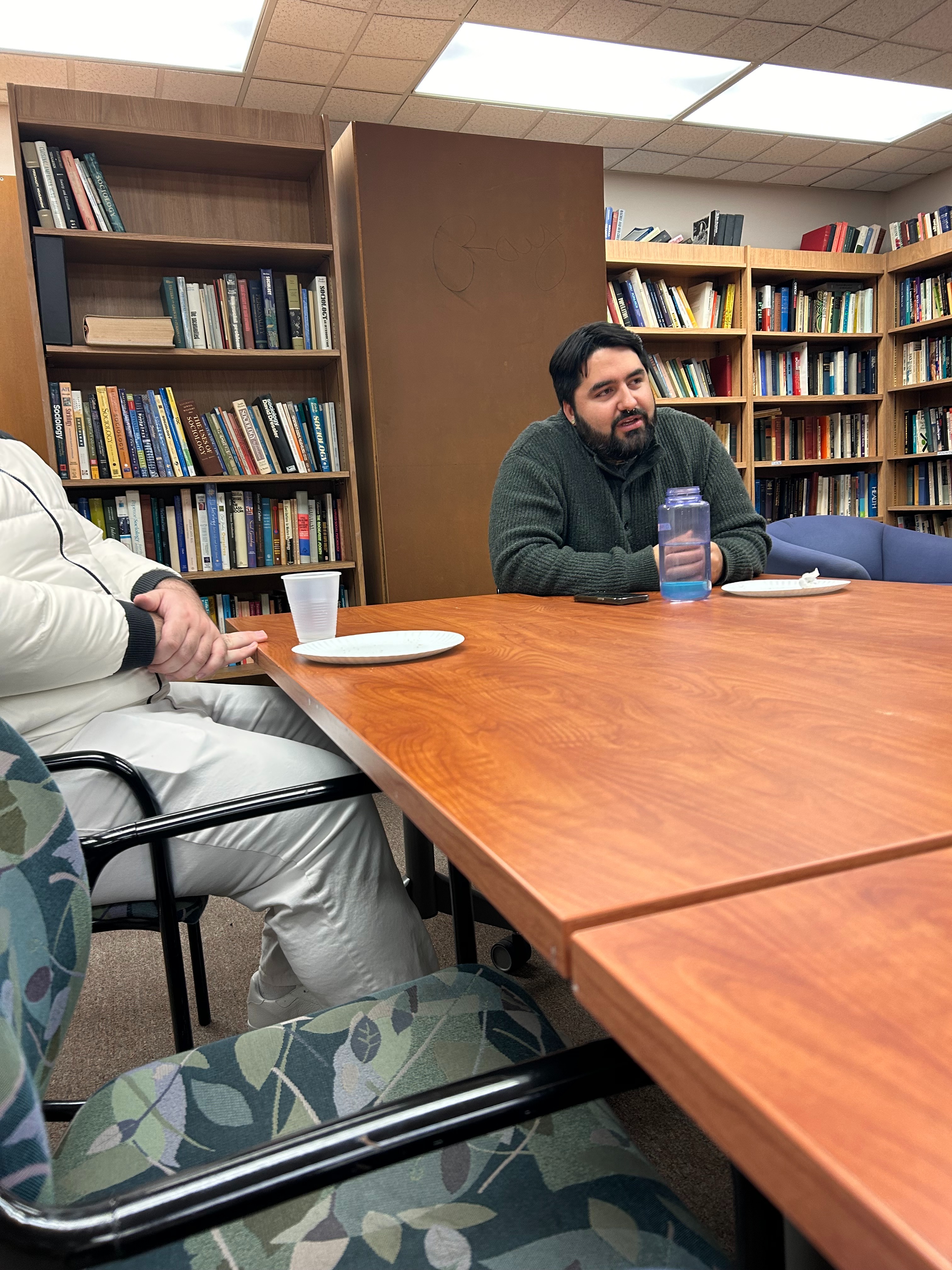 Conference room with bookshelves, a wooden table, and two people seated, one holding a blue water bottle.